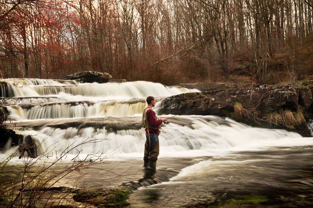man in waders and fishing vest standing next to waterfalls fishing